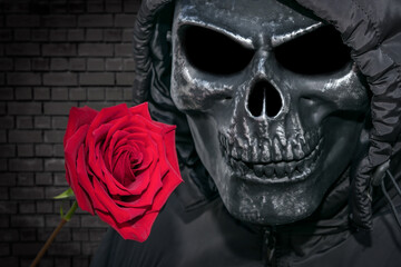 Ominous form of death with a red rose on a brick wall background. Man in a skull mask and a flower...