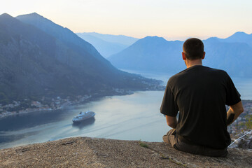 Fototapeta na wymiar A man in a black T-shirt sits looking down on a cruise ship in a sea bay. Against the backdrop of a mountain. Pre-sunset time. The concept of tourism in Montenegro.