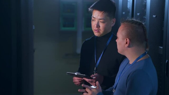 Multi-ethnic team of engineers talking and checking server racks in big data center spbas. Close-up view of two young men discuss business and hold tablet in hand, control complex systems and networks