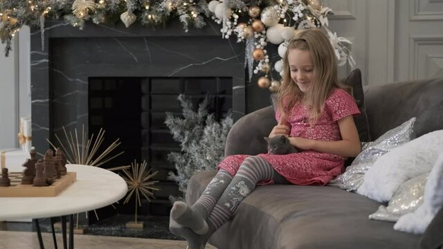 Smiling Little Girl Stroking Cute Kitten while Sitting on the Couch in Xmas Day Indoors. Slow Motion. Friendship Between Human and Pet. Christmas and New Year Celebration Concept