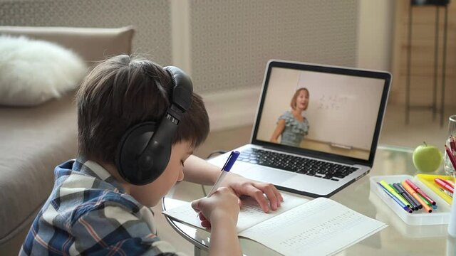 Online home school for junior schoolchildren. Little boy in checkered shirt solves sums with teacher on screen at maths lesson spbas spbd via notebook at table in room