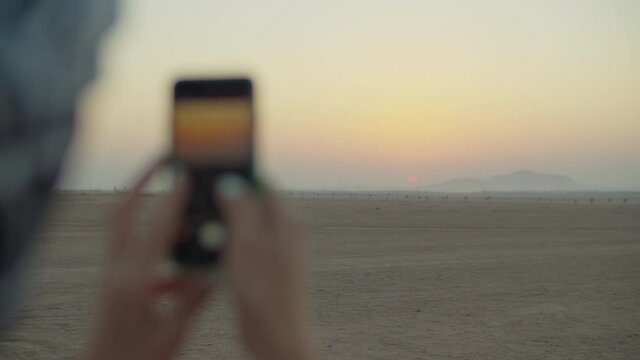 Female hands taking picture of sunrise on mobile phone in the desert. Tourist enjoy desert dawn. Traveler taking photo of beautiful desert sunrise.
