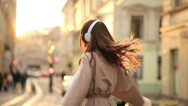 Young adult happy woman dancing in downtown very emotional. Active happy girl in wireless headphones jumping, dancing and having fun outdoors in city street at sunset