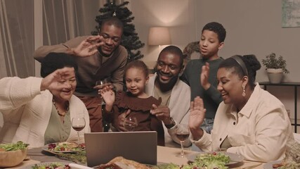 Medium long of happy African American family members of different generations sitting at festive table with foods and drinks on, video calling via portable computer on Christmas eve - Powered by Adobe