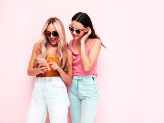 Two young beautiful smiling brunette hipster female in trendy summer clothes. Sexy carefree women posing near pink wall. Positive models having fun. Cheerful and happy. Holding smartphone. Using apps