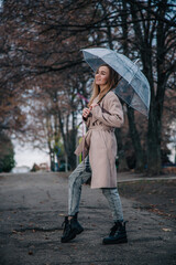 A young, slender, beautiful girl in a coat walks in the rain with a transparent umbrella in the park