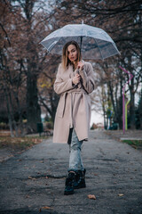 A young, slender, beautiful girl in a coat walks in the rain with a transparent umbrella in the park