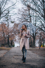 A young, beautiful girl with a disposable cup of coffee walks in the park under a transparent umbrella in the rain