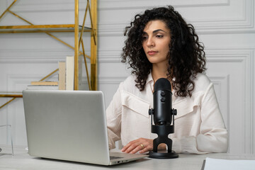 Curly-haired brunette woman with a microphone and laptop, talking and recording a podcast in the...