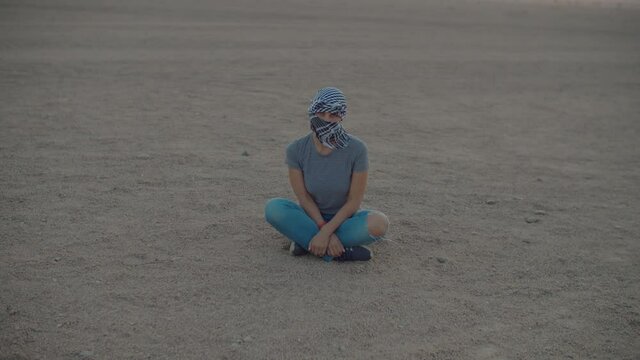 Woman in checkered keffiyeh sitting in the desert looking at camera. Caucasian female tourist sitting on desert sand.
