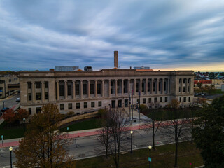 Aerial view of Kenosha County Courthouse. Park and trees across street. Checkered blue sky.  Bricks lining sidewalk. Sidewalk lights glowing.  Chimney stack seen on roof of building.  