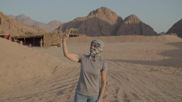 Tourist in checkered keffiyeh trying to find mobile network using cell phone standing in desert. Woman searching for mobile signal in desert. No cell operator signal.