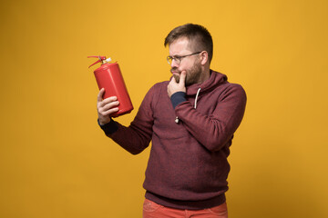 Puzzled man with glasses holds a fire extinguisher in hand and looks at it thoughtfully. Yellow...