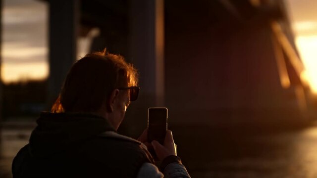 A man on the background of sunset looks at the phone and photographs a beautiful panorama. Mobile lifestyle in the modern world. Shooting sunsent on a smartphone