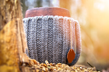 Clay mug in woolen knitted cover. Steam from the mug on the background of autumn forest and sunlight.