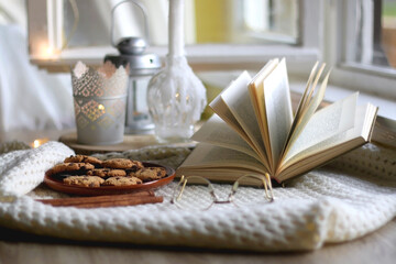 Plate of chocolate chip cookies, cinnamon sticks, soft blanket, open book and lit candles. Hygge at home. Selective focus.
