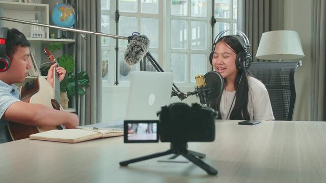 Camera Recording, Asian Kid Girl In Headphones Singing Into Microphone While Boy Playing The Guitar During Online Livestream In Podcast Studio
