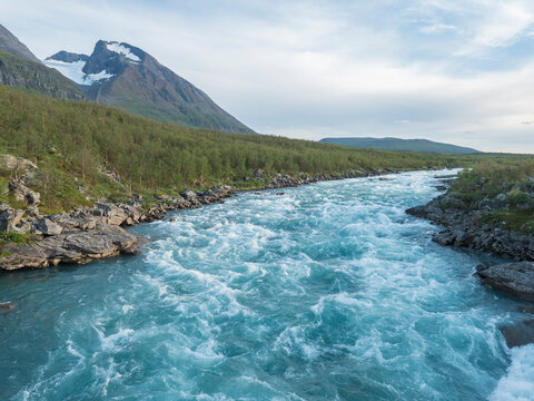 Blue wild water of Vuojatadno river rapids with mount Akka, Ahkka massif with snow and glacier and birch tree forest. Beatiful northern artic landscape in Swedish Lapland. Summer evening