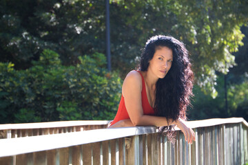 beautiful adult woman, brunette with curly hair and latina is leaning on the wooden railing of a pond in the park while looking at the camera making different expressions. Holiday and travel concept