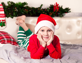 Boy kid in funny Santa cap and Christmas pajamas lie on bed at home