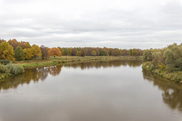 Autumn landscape with a river. Cloudy autumn day by the river. View of the river with trees and bushes on the bank. Bright colors of autumn on a cloudy day.