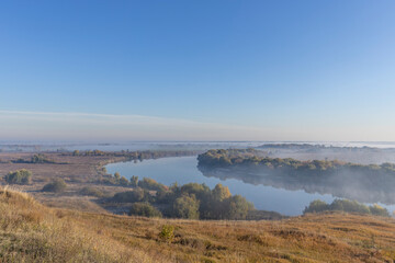 Autumn landscape in the early morning overlooking the river. A wide river and endless expanses of fields. Yellow leaves on trees and bushes are illuminated by the rays of the rising sun.