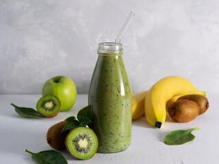 A fresh smoothie made of kiwi, banana, spinach leaves and apple juice in a glass bottle on a light table. Yellow bananas, kiwi, spinach leaves and a green apple in the background