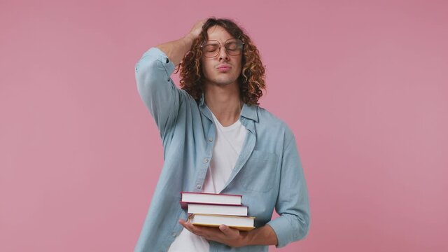 Confused pensive young curly man 20s wears blue shirt white t-shirt wearing translucent pink glasses hold a pile of books spreading hand isolated on pastel plain light pink background studio portrait