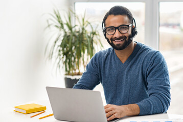 Portrait of cheerful Indian guy in wireless headset using laptop computer sitting in contemporary loft style office space, Hispanic man is making video call, talking online on distance