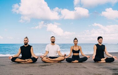 Group of diverse male and female friends have together morning meditation at coastline beach breathing during holistic healing, people in sportswear exercising in lotus yoga pose feeling calmness