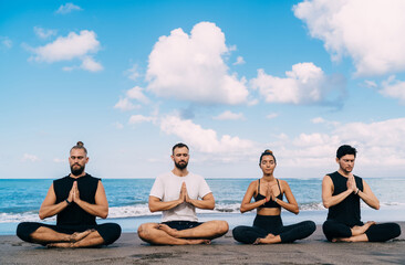 Young male and female in lotus pose holding hands in namaste enjoying spiritual meditation at coastline beach, group of friends have together morning yoga practice at seashore in Indonesia