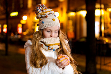 Night portrait of beautiful girl in knitted hat and scarf holding garland in her palms, smiling, enjoying winter outdoors. Winter joys. Winter vacation. Positive emotions. Happiness