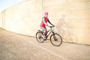Naklejka premium Young caucasian active woman in warm pink fitness clothes riding bike outdoors near wall on streets or athletic sports ground on winter or spring early morning sunrise. Active lifestyle, transport