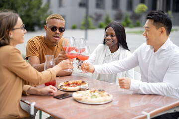 Multiracial business team having lunch with wine and pizza at outdoor cafe. Concept of teambuilding and corporate event. Idea of rest and leisure on job. People sitting at wooden table and talking