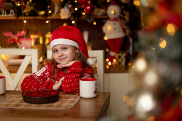 Fototapeta na wymiar a cute little girl child in a red sweater and a Santa Claus hat is in the kitchen drinking tea with a pie and waiting for the new year or Christmas