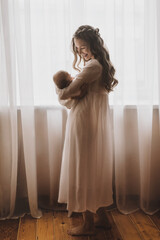 Fototapeta premium Beautiful young mother holds newborn baby girl in white lighter room at home. Tenderness cozy atmosphere