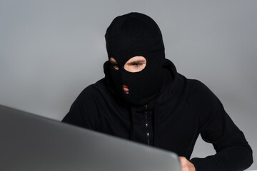 Hacker in balaclava looking at computer monitor isolated on grey