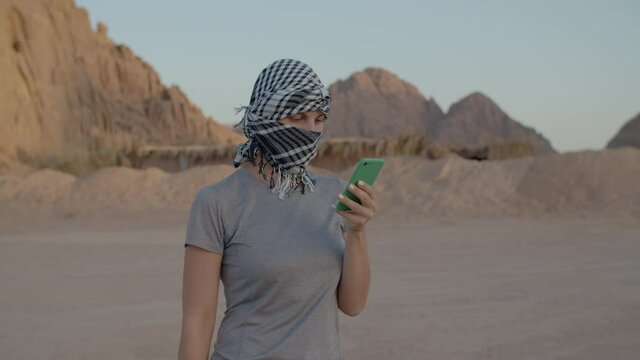 Female tourist in checkered keffiyeh trying to find mobile network using smart phone standing in desert. Woman searching for mobile signal to make a call in desert. 