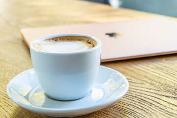 White big cup of cappuccino with foam on table. Morning coffee concept with stylish toning and place for text. A cup of coffee on the table near the laptop. 
