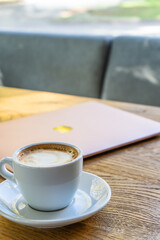 White big cup of cappuccino with foam on table. Morning coffee concept with stylish toning and place for text. A cup of coffee on the table near the laptop. Online classes, training, and blogging
