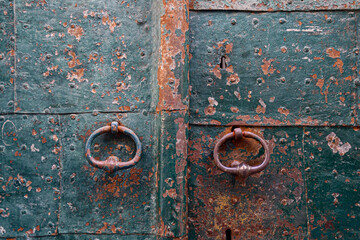 Detail of a weathered, rusty metal door with a pair of knockers, peeled green paint and studs, Alassio, Savona, Liguria, Italy