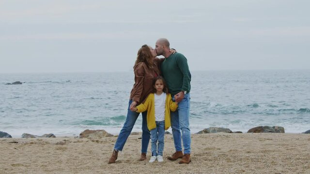 Young happy family is spending time on ocean beach, parents are kissing each other, holding their daughter’s arms, looking at camera, live in harmony and love, Slow motion. 