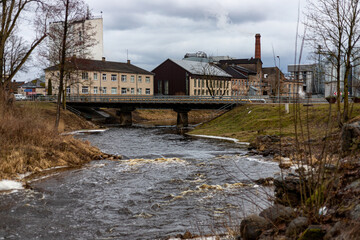 Fototapeta na wymiar transport bridge over river Iecava in Latvia. Big rapid massive stream of river in spring after ice meltdown, Living house and old factory in distance
