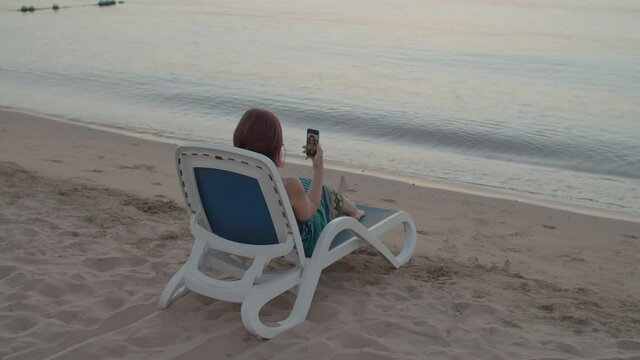 Woman taking selfie picture sitting by the sea using cell phone. Over the shoulder back view of woman taking selfie photo memories on mobile phone sitting on beach chair.