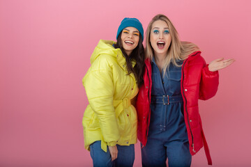 two attractive girl friends active women posing on pink background in colorful winter down jacket...
