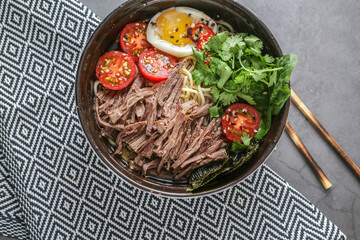 a plate of delicious beef soup, pasta with tomato, half a fried egg, sesame seeds and herbs on a gray background with wooden Chinese sticks.