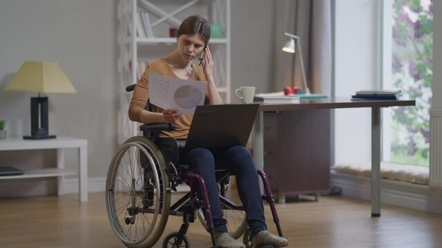 Focused genius Caucasian disabled woman in wheelchair talking on phone sitting with laptop and paperwork in home office. Portrait of intelligent freelancer discussing idea in slow motion indoors