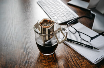 Convenient paper drip coffee bag in glass cup on dark office table. Making freshly brewed coffee at work