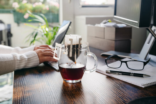 Glass Cup Of Freshly Brewed Coffee With Convenient Drip Coffee Bag On Dark Office Table. Womans Hands Typing On Keboard At The Background