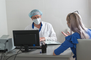 Obraz premium A doctor in a white coat, mask and cap is sitting at table with a patient in the office and discussing the treatment protocol. Consultative reception